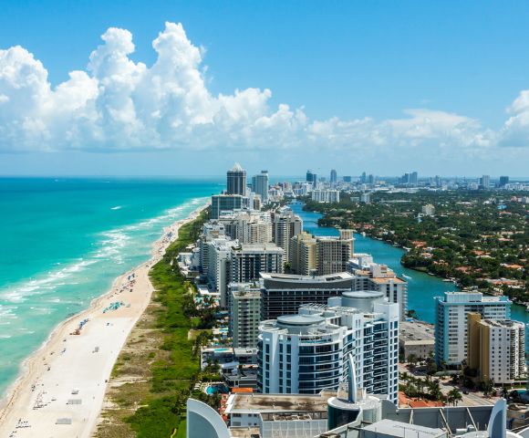 Aerial view of Miami Beach, showcasing turquoise ocean waves, sandy shoreline, and high-rise buildings under a bright blue sky with fluffy clouds.