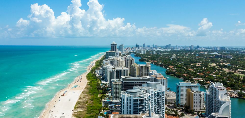 Aerial view of Miami Beach, showcasing turquoise ocean waves, sandy shoreline, and high-rise buildings under a bright blue sky with fluffy clouds.