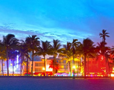Tropical beach at dusk with silhouetted palm trees and brightly lit neon buildings in vibrant colors, creating a lively and energetic atmosphere.