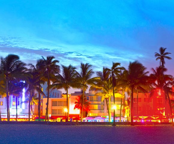 Tropical beach at dusk with silhouetted palm trees and brightly lit neon buildings in vibrant colors, creating a lively and energetic atmosphere.