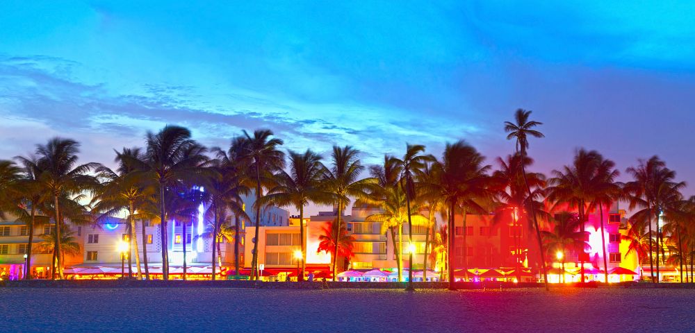 Tropical beach at dusk with silhouetted palm trees and brightly lit neon buildings in vibrant colors, creating a lively and energetic atmosphere.