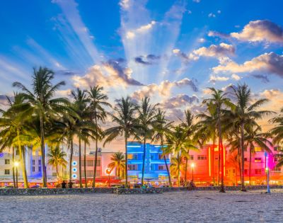 Palm trees silhouette against a vibrant sunset sky, with colorful Art Deco buildings lit in pink and blue. The scene is lively and tropical.