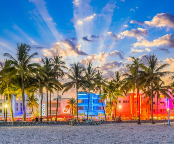 Palm trees silhouette against a vibrant sunset sky, with colorful Art Deco buildings lit in pink and blue. The scene is lively and tropical.