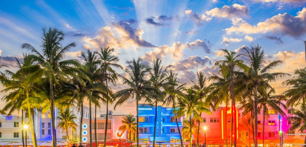 Palm trees silhouette against a vibrant sunset sky, with colorful Art Deco buildings lit in pink and blue. The scene is lively and tropical.
