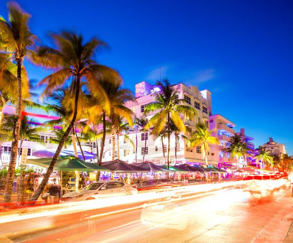Vibrant street scene at dusk with neon-lit Art Deco buildings, palm trees, and motion-blurred car lights, creating a lively, energetic atmosphere.
