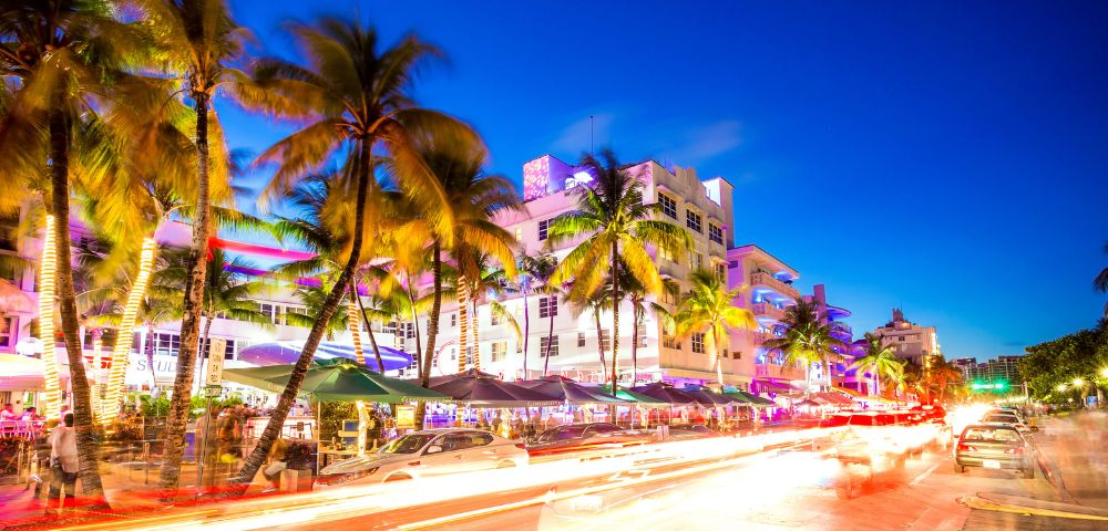 Vibrant street scene at dusk with neon-lit Art Deco buildings, palm trees, and motion-blurred car lights, creating a lively, energetic atmosphere.