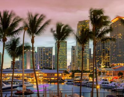 Skyline at dusk with illuminated skyscrapers, palm trees, and boats at a marina. The sky is a blend of pink and purple hues, creating a vibrant cityscape.