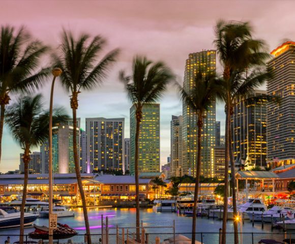 Skyline at dusk with illuminated skyscrapers, palm trees, and boats at a marina. The sky is a blend of pink and purple hues, creating a vibrant cityscape.