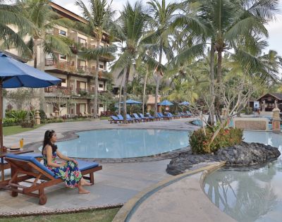 Woman in floral dress relaxes on a lounge chair by a resort pool with blue umbrellas and palm trees. Tropical setting, tranquil atmosphere.