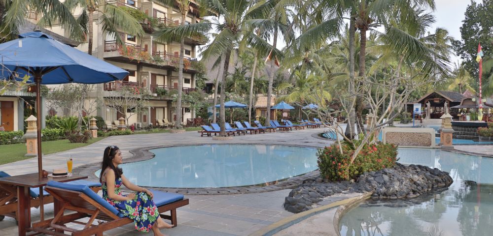 Woman in floral dress relaxes on a lounge chair by a resort pool with blue umbrellas and palm trees. Tropical setting, tranquil atmosphere.