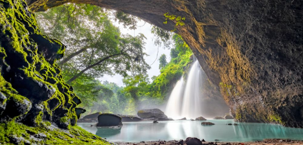 A cascading waterfall and tranquil turquoise pool viewed from inside a serene cave with green, moss-covered rock in Khao Yai National Park.