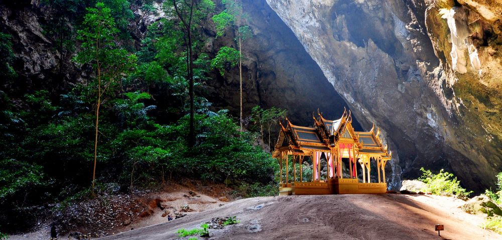 A four-gabled pavilion with a distinctive roof nestled in the large dome of Phraya Nakhon cave, illuminated by dappled sunlight.