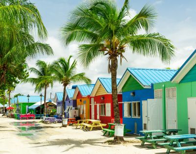 A row of vibrant beach huts in red, blue, yellow, and green with blue roofs line a sandy path. Palm trees add a tropical feel under a partly cloudy sky.