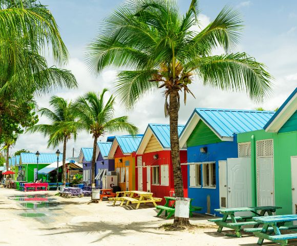 A row of vibrant beach huts in red, blue, yellow, and green with blue roofs line a sandy path. Palm trees add a tropical feel under a partly cloudy sky.