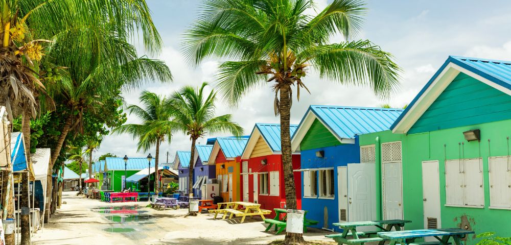 A row of vibrant beach huts in red, blue, yellow, and green with blue roofs line a sandy path. Palm trees add a tropical feel under a partly cloudy sky.