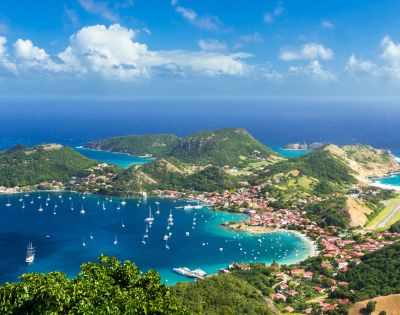 Aerial view of a vibrant coastal town with red-roofed buildings, lush green hills, and a bay filled with sailing boats under a bright blue sky with fluffy clouds.