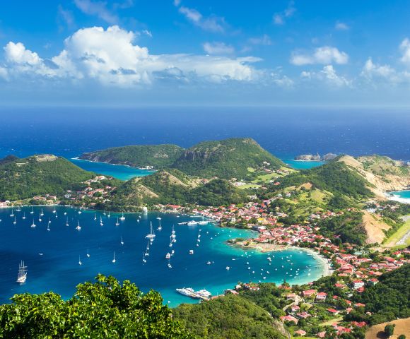 Aerial view of a vibrant coastal town with red-roofed buildings, lush green hills, and a bay filled with sailing boats under a bright blue sky with fluffy clouds.
