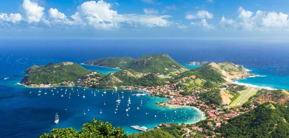 Aerial view of a vibrant coastal town with red-roofed buildings, lush green hills, and a bay filled with sailing boats under a bright blue sky with fluffy clouds.