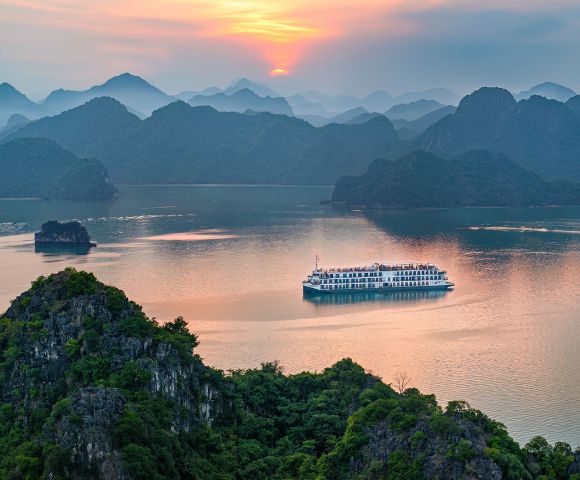 A cruise ship sails on a serene bay surrounded by lush green islands, under a sunset sky with warm, pink hues. The atmosphere is tranquil and picturesque.