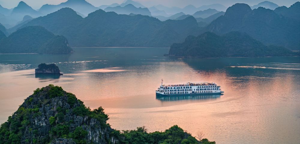 A cruise ship sails on a serene bay surrounded by lush green islands, under a sunset sky with warm, pink hues. The atmosphere is tranquil and picturesque.