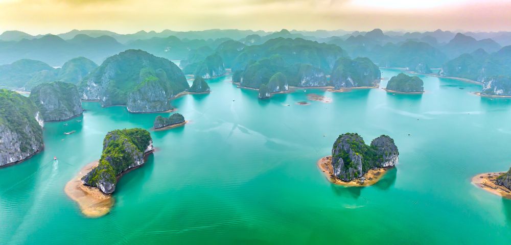 Aerial view of Ha Long Bay, Vietnam, showcasing emerald waters dotted with lush, limestone islands under a hazy sky, evoking tranquility and grandeur.