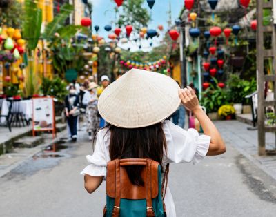 A woman with a conical hat and backpack walks through a vibrant street adorned with colorful lanterns, evoking a lively, festive atmosphere.