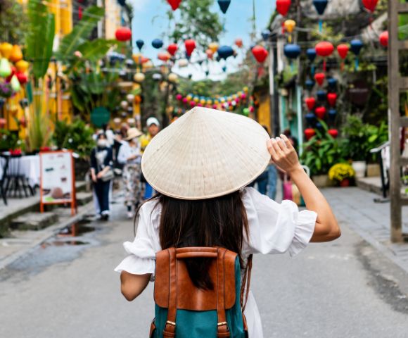 A woman with a conical hat and backpack walks through a vibrant street adorned with colorful lanterns, evoking a lively, festive atmosphere.