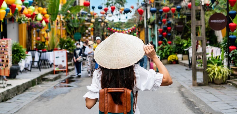 A woman with a conical hat and backpack walks through a vibrant street adorned with colorful lanterns, evoking a lively, festive atmosphere.