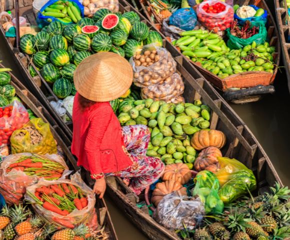 Aerial view of a vibrant floating market with boats filled with colorful produce, including watermelons, pumpkins, and pineapples. A person in a straw hat sits on one boat, conveying a bustling, lively scene.
