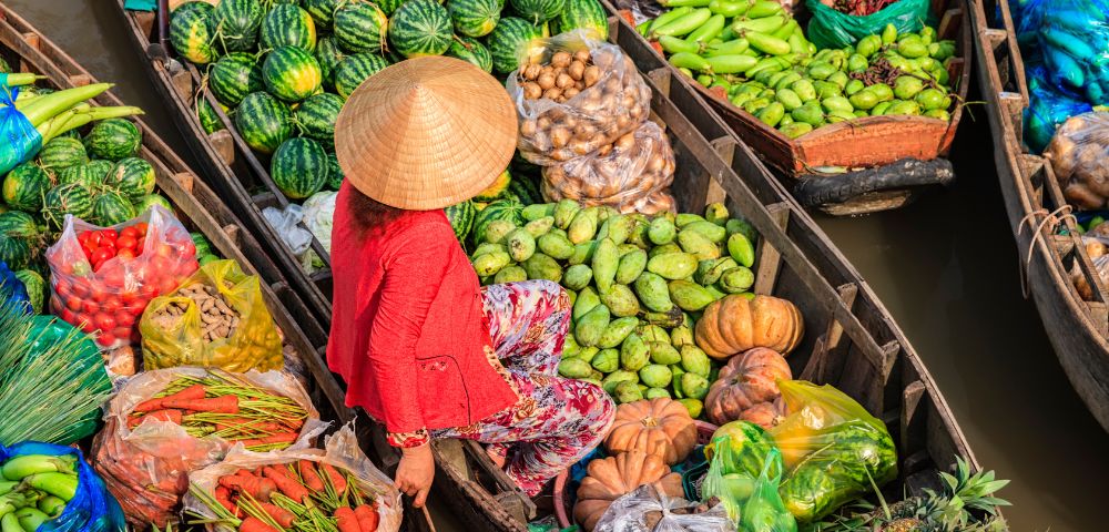 Aerial view of a vibrant floating market with boats filled with colorful produce, including watermelons, pumpkins, and pineapples. A person in a straw hat sits on one boat, conveying a bustling, lively scene.