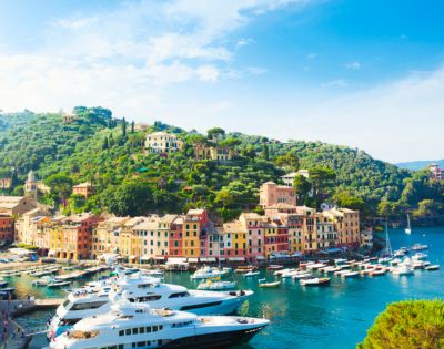 Picturesque coastal town with vibrant pastel buildings nestled on a green hillside. Luxurious yachts float on the serene, blue waters under a clear sky.