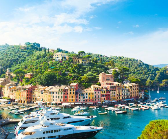 Picturesque coastal town with vibrant pastel buildings nestled on a green hillside. Luxurious yachts float on the serene, blue waters under a clear sky.