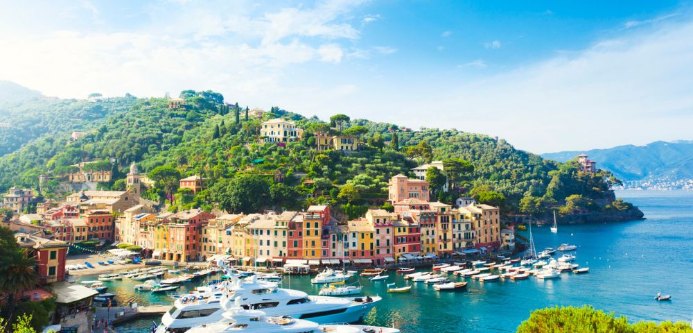 Picturesque coastal town with vibrant pastel buildings nestled on a green hillside. Luxurious yachts float on the serene, blue waters under a clear sky.