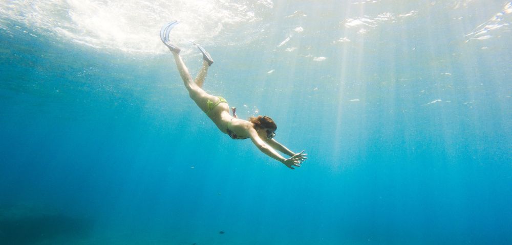 A person in a swimsuit dives gracefully underwater in clear blue ocean, with sunlight streaming down, creating a serene and adventurous atmosphere.