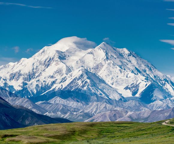 Snow-capped mountain under clear blue sky, with lush green hills in the foreground. The scene conveys tranquility and natural beauty.