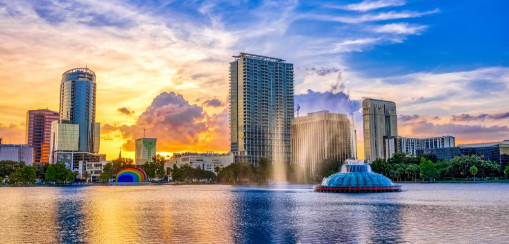 Sunset over a cityscape with tall buildings reflecting in a calm lake. A fountain sprays water, and colorful clouds fill the sky, creating a serene ambiance.