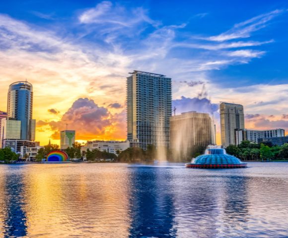 Sunset over a cityscape with tall buildings reflecting in a calm lake. A fountain sprays water, and colorful clouds fill the sky, creating a serene ambiance.