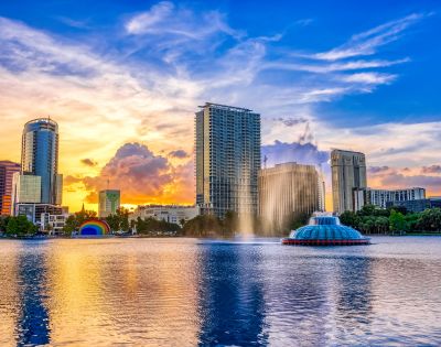 Sunset over a cityscape with tall buildings reflecting in a calm lake. A fountain sprays water, and colorful clouds fill the sky, creating a serene ambiance.