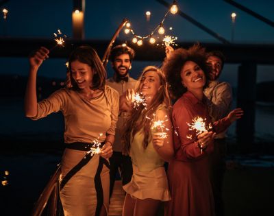 Five people smiling and holding sparklers on a bridge at night, with string lights above them, creating a festive and joyful atmosphere.
