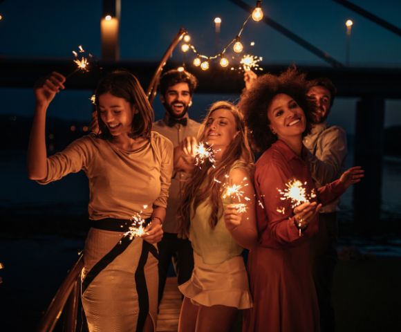 Five people smiling and holding sparklers on a bridge at night, with string lights above them, creating a festive and joyful atmosphere.