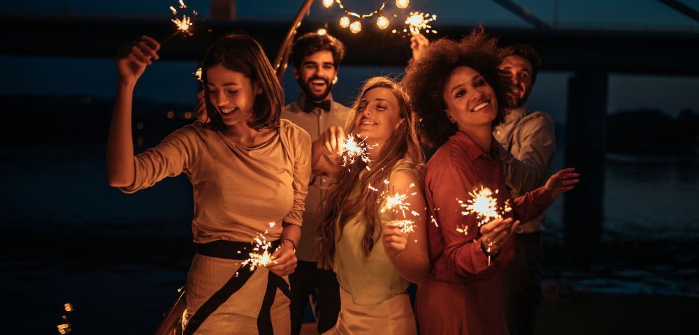 Five people smiling and holding sparklers on a bridge at night, with string lights above them, creating a festive and joyful atmosphere.