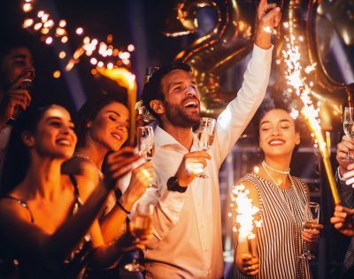 A joyful group celebrates with sparklers and champagne, surrounded by golden balloons. They appear excited, creating a festive and energetic atmosphere.