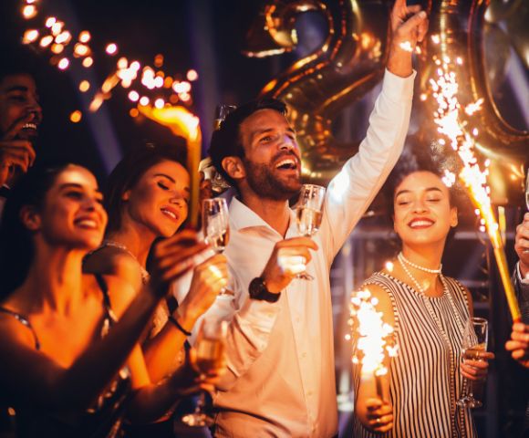 A joyful group celebrates with sparklers and champagne, surrounded by golden balloons. They appear excited, creating a festive and energetic atmosphere.