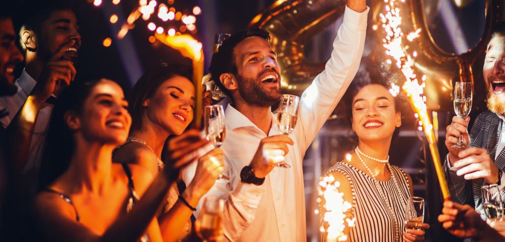 A joyful group celebrates with sparklers and champagne, surrounded by golden balloons. They appear excited, creating a festive and energetic atmosphere.