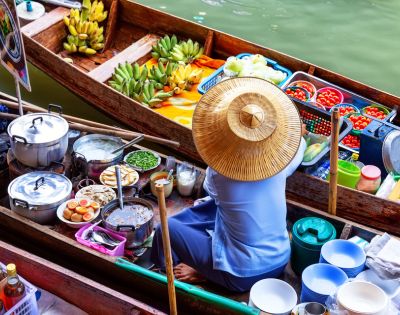 A vendor in a straw hat sits in a colorful floating market boat, surrounded by fresh produce, steaming pots, and vibrant baskets of fruit on a calm river.