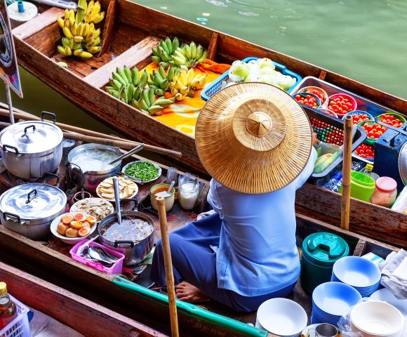 A vendor in a straw hat sits in a colorful floating market boat, surrounded by fresh produce, steaming pots, and vibrant baskets of fruit on a calm river.