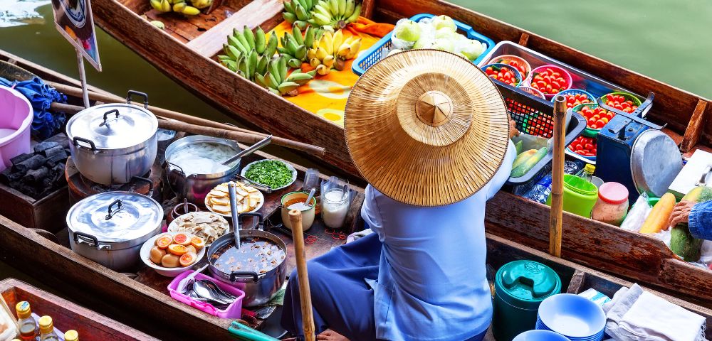 A vendor in a straw hat sits in a colorful floating market boat, surrounded by fresh produce, steaming pots, and vibrant baskets of fruit on a calm river.