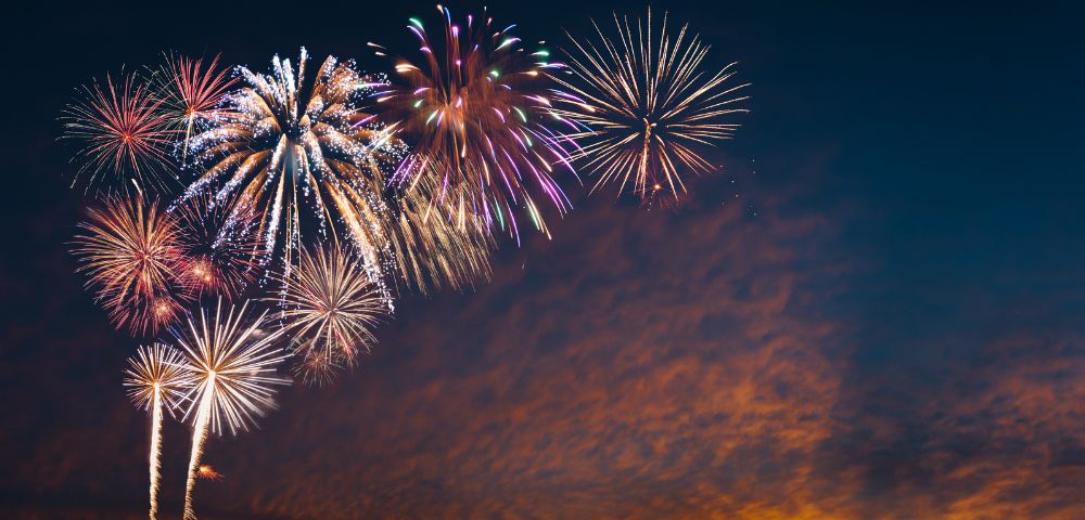 A vibrant fireworks display illuminates the night sky with colorful bursts, set against a serene backdrop of twilight clouds, evoking a festive atmosphere.