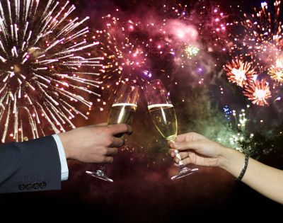 Two hands toasting with champagne glasses against a vibrant backdrop of colorful fireworks, creating a festive and celebratory atmosphere.