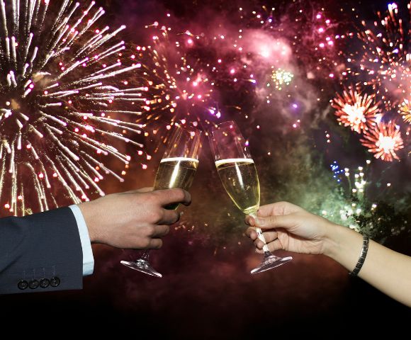 Two hands toasting with champagne glasses against a vibrant backdrop of colorful fireworks, creating a festive and celebratory atmosphere.